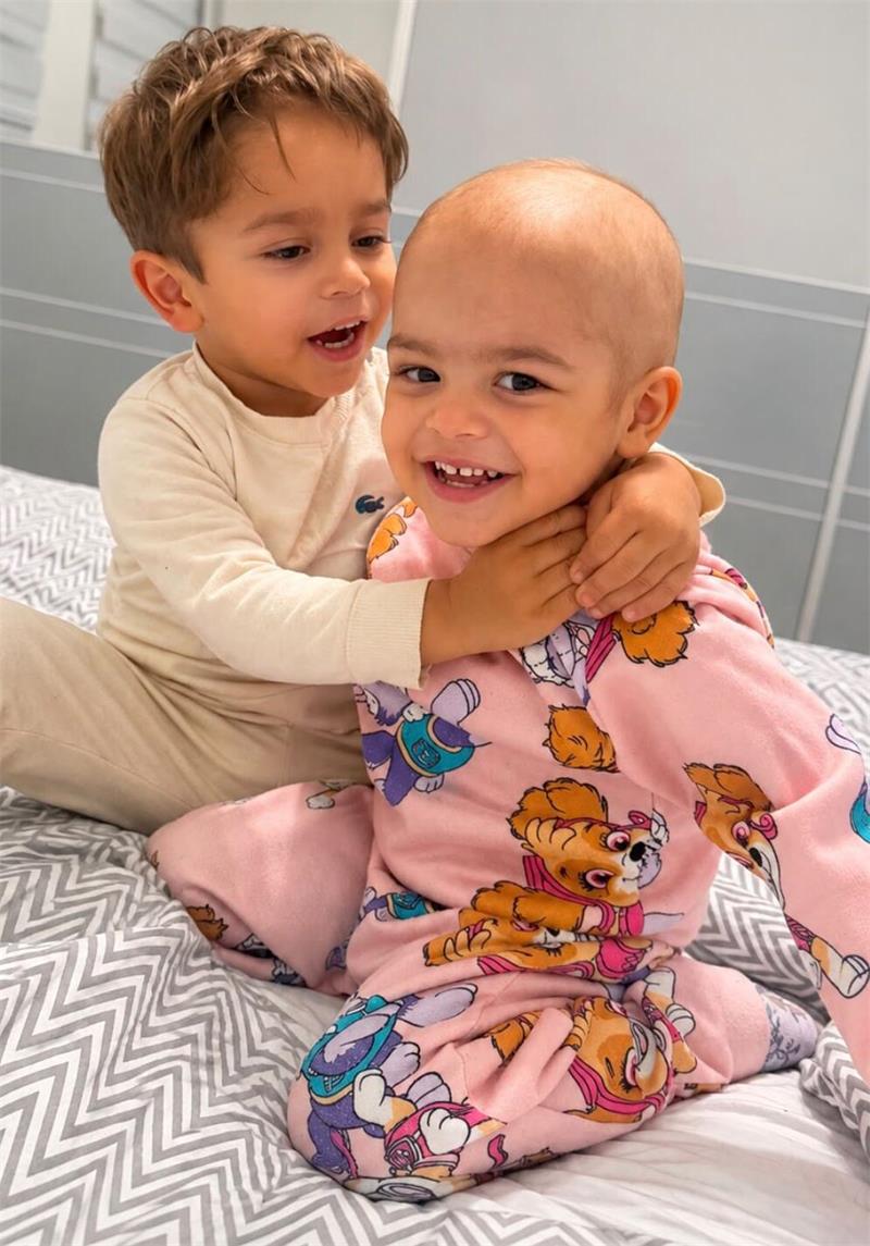 Zwei Kinder sitzen auf einem Bett, das eine Kind umarmt das andere, beide lächeln, das umarmte Kind trägt einen Schlafanzug mit buntem Bärchenmuster