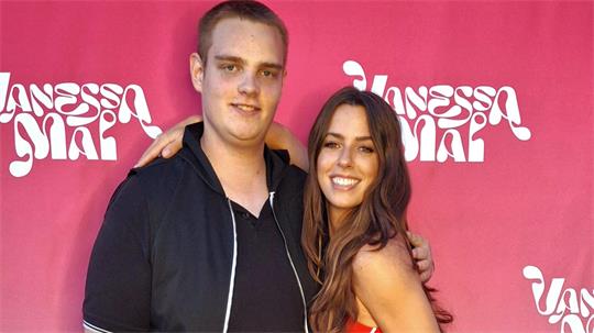 Lukas Tscherner und Schlagersängerin Vanessa Mai stehen vor einem pinken Hintergrund mit weißer Schrift und umarmen sich lächelnd.