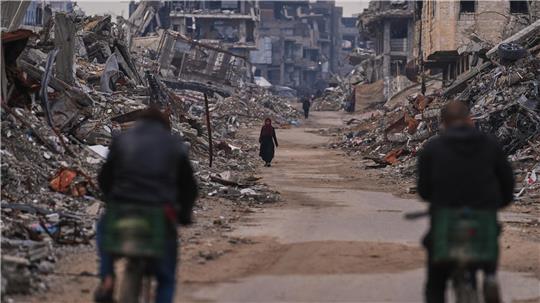 Zwei Personen auf Fahrrädern fahren auf einer von Trümmern gesäumten Straße, eine weitere Person geht in der Ferne.