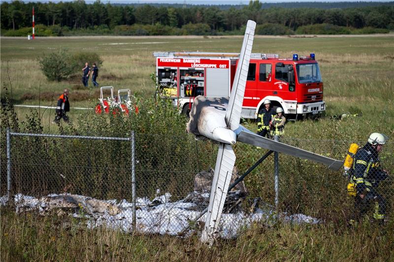 Kleinflugzeug stürzt in Bamberg ab - Pilot schwer verletzt Trümmerteile eines Flugzeugs hängen in einem Zaun, während Einsatzkräfte der Feuerwehr an der Absturzstelle stehen.