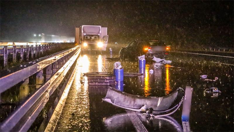 Nasse Autobahn bei Nacht mit Unfallfahrzeug, Trümmern und einem Lkw mit eingeschalteten Scheinwerfern im Hintergrund