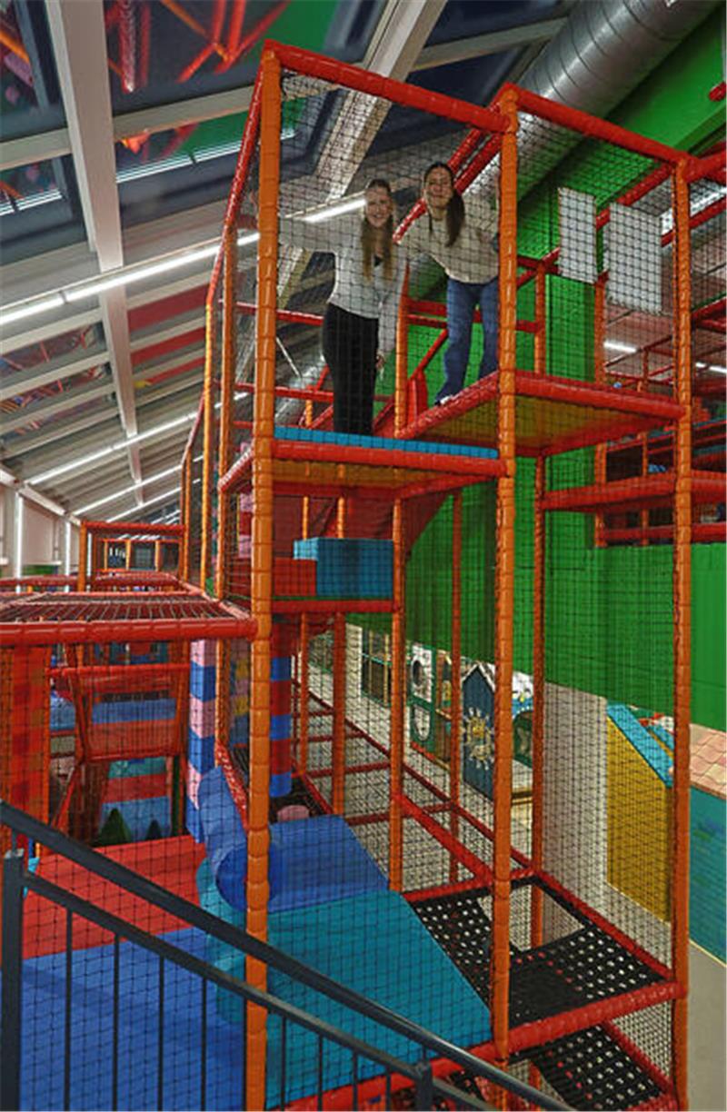 Zwei Personen stehen auf einer erhöhten Plattform eines roten Klettergerüsts in einer Halle mit grüner Wand und Glasdach