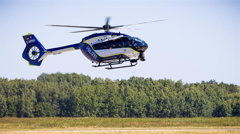 Polizeihubschrauber in Blau und Weiß schwebt über einer Grasfläche vor einem Wald.
