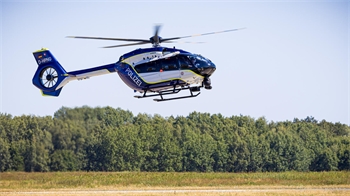 Polizeihubschrauber in Blau und Weiß schwebt über einer Grasfläche vor einem Wald.