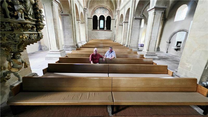 Zwei ältere Männer sitzen in einer großen Kirche auf einer der vorderen Holzbänke mit hohen Rundbögen und verzierten Säulen im Hintergrund. 