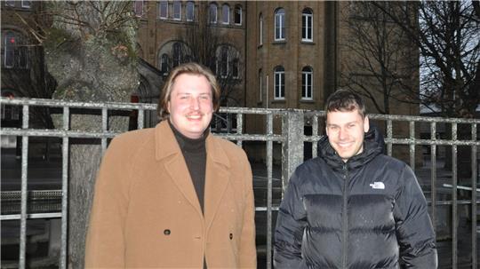 Zwei Männer stehen vor einem historischen Gebäude mit Rundturm und mehreren Fenstern, umgeben von Bäumen und einem Zaun.