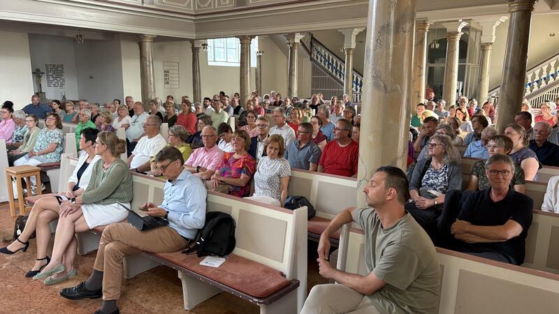 Viele Menschen sitzen in einer Kirche auf Holzbänken und hören aufmerksam zu