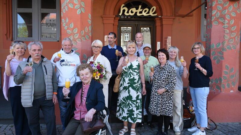 Eine Gruppe an Menschen steht vor einem Hotel und trinkt Sekt.