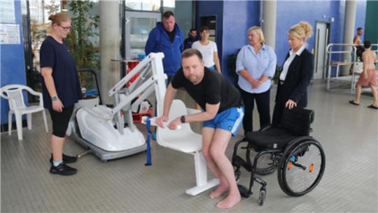 Mehrere Personen stehen und sitzen um einen beweglichenLift, eine Person in Sportkleidung steigt vom Rollstuhl in den Lift-Sitz um.