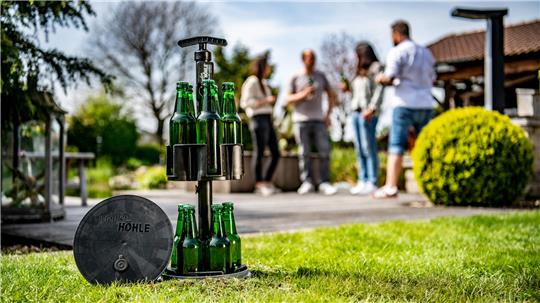 Die ausgefahrene Hopfenhöhle, befüllt mit grünen Glasflaschen, ragt aus dem Erdreich. Im Hintergrund stehen vier Personen.