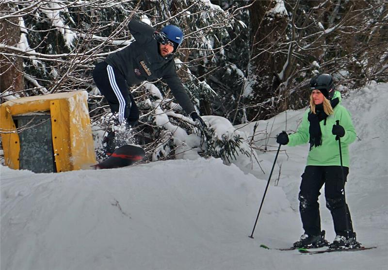 Eine Person in schwarzer Kleidung springt mit Snowboard über eine Schneehügelkante, daneben steht eine Person in grüner Jacke mit Skiern und Skistöcken.