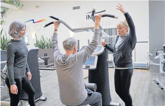 „Und noch einmal, kräftig.“ Elke Paschek (r.) mit Eberhard Kleuker, erkrankt an einem Tumor im Rachenraum, im Braunschweiger Injoy-Fitnessstudio. Kurs-Trainerin Jana Cordes (li.) beobachtet beide bei der Übung am sogenannten „Latzug“, der große Muskelpartien am Rücken trainiert. Foto: Stefan Lohmann/Regios24