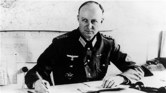 Ein Mann in Uniform mit Abzeichen sitzt an einem Schreibtisch und hält einen Stift in der rechten Hand.