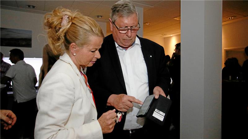 Zwei Personen in formeller Kleidung stehen in einem Innenraum, eine hält ein schwarzes Handy, die andere trägt einen weißen Blazer und hat eine Hochsteckfrisur.