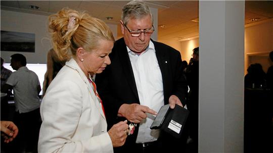 Zwei Personen in formeller Kleidung stehen in einem Innenraum, eine hält ein schwarzes Handy, die andere trägt einen weißen Blazer und hat eine Hochsteckfrisur.