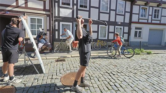 Mehrere Personen auf einem gepflasterten Platz vor einem Fachwerkhaus, eine Person hebt ein Mikrophon über den Kopf, andere bedienen Kamera und Fahrrad.