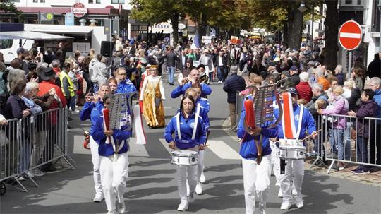 Eine Musikkapelle zieht eine Straße entlang. Rechts und links befinden sich Zuschauer.