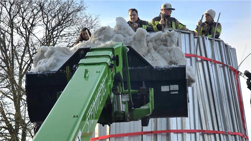 Drei Personen stehen auf einer großen Metallstruktur, davor ein grüner Teleskoplader mit einer Schaufel voller Schnee oder Eis.