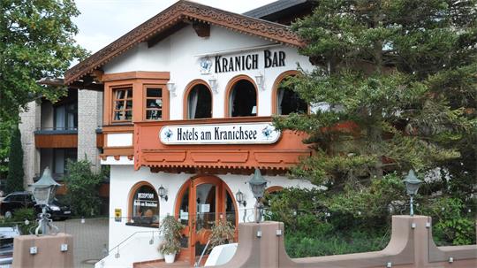 Fachwerkhaus mit dem Schriftzug 'KRANICH BAR' und 'Hotels am Kranichsee' an der Fassade, umgeben von Bäumen und einem Zaun im Vordergrund