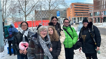 Gruppe von sechs Personen mit Winterkleidung und Rucksäcken steht auf schneebedecktem Bürgersteig in urbaner Umgebung mit Gebäuden und Bäumen im Hintergrund.