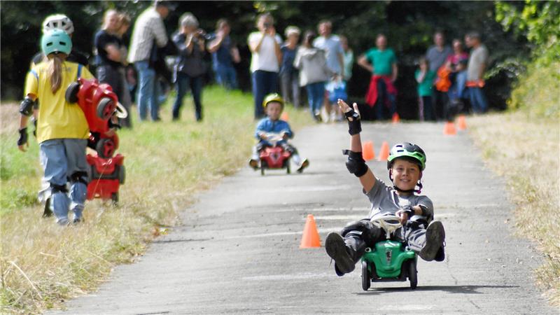 Kind sitzt auf grünem Tretfahrzeug auf asphaltierter Strecke, hebt eine Hand, umgeben von weiteren Kindern und Erwachsenen am Wegesrand.