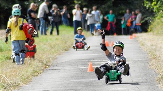 Kind sitzt auf grünem Tretfahrzeug auf asphaltierter Strecke, hebt eine Hand, umgeben von weiteren Kindern und Erwachsenen am Wegesrand.