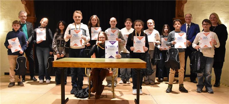 Gruppe von Kindern und Erwachsenen steht in einer Reihe und hält Urkunden in der Hand, vor einem Holztisch in einem Raum mit heller Wand.