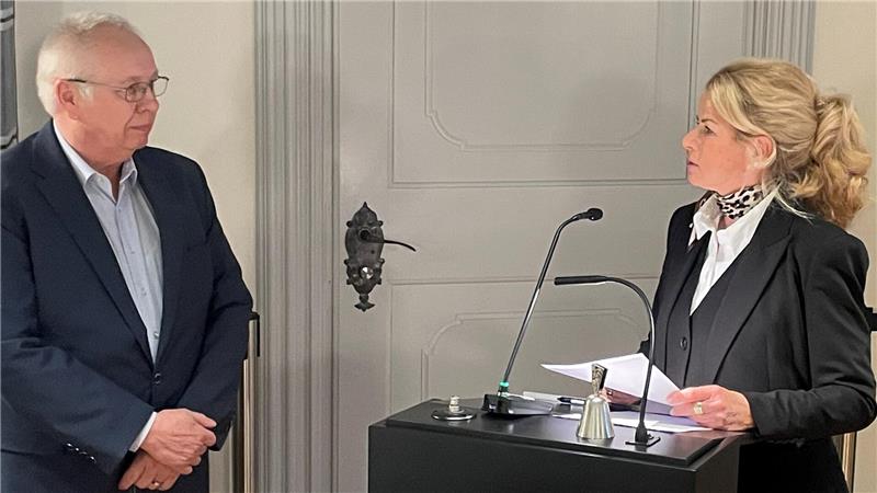 Frau in schwarzem Anzug mit weißem Hemd steht an einem schwarzen Rednerpult und hält Papiere, Mann in dunklem Sakko und Jeans steht daneben vor einer hellen Tür.