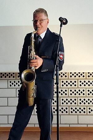 Person in Polizeiuniform spielt Saxophon vor Mikrofonständer in Innenraum mit perforierter Wandverkleidung.