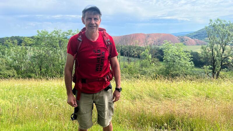 Mann mit rotem T-Shirt, grauer Kappe und Rucksack steht auf einer Wiese vor grünen Bäumen und einem roten Hügel im Hintergrund.