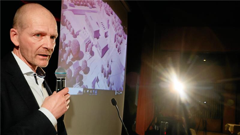 Mann mit Mikrofon vor einer Leinwand mit einem Stadtmodell, im Hintergrund starke Lichtquelle