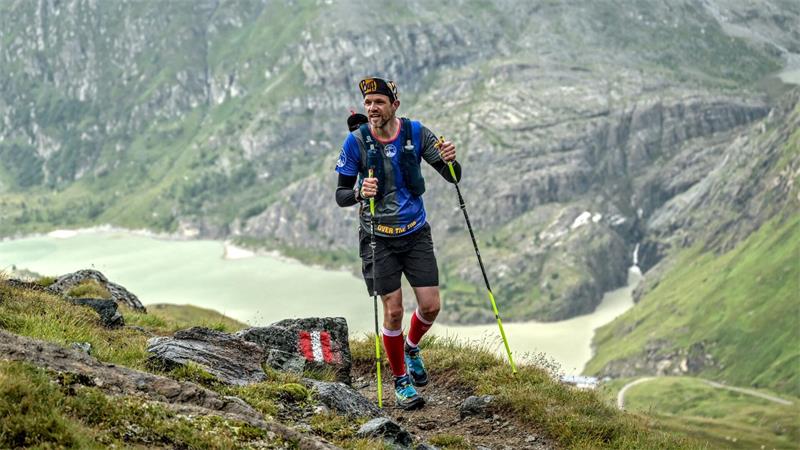 Mann mit Trekkingstöcken läuft auf einem Bergpfad mit Fels und Gras vor einer Berglandschaft mit See