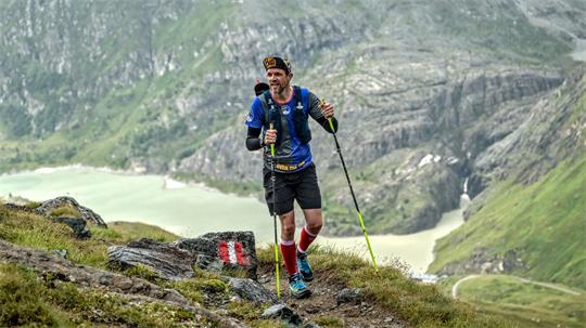 Mann mit Trekkingstöcken läuft auf einem Bergpfad mit Fels und Gras vor einer Berglandschaft mit See