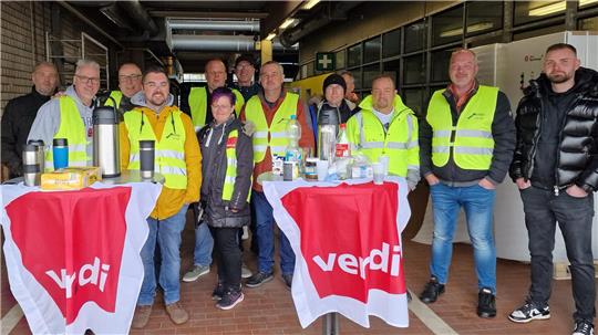 Gruppe von Menschen in Warnwesten steht in einem überdachten Bereich neben Tischen mit roten Tüchern und Thermoskannen.