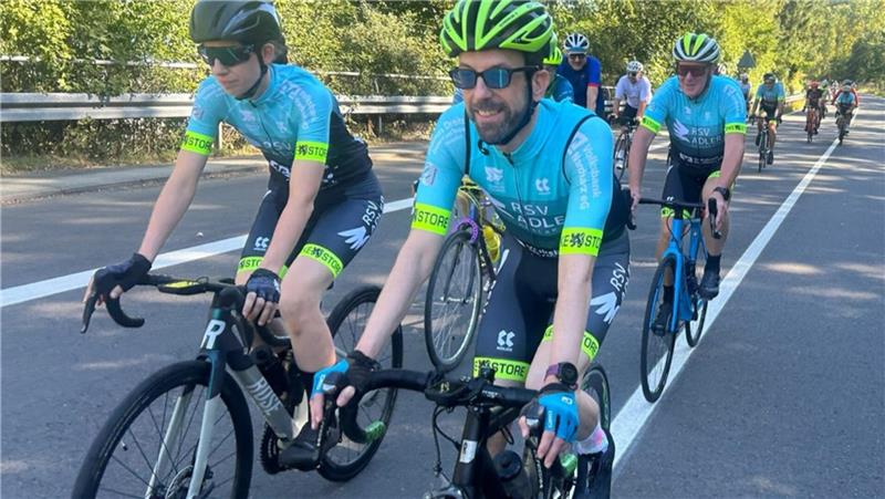 Mehrere Radrennfahrer in blauen Trikots und gelb-schwarzen Helmen fahren auf einer Straße in einer Gruppe