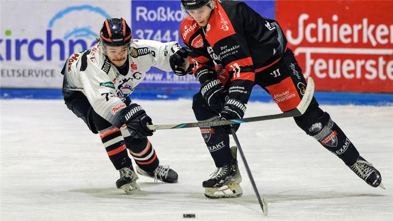 Zwei Eishockeyspieler in Aktion, einer in weißem Trikot, der andere in schwarzem Trikot mit roten Akzenten, kämpfen um den Puck auf dem Eis.