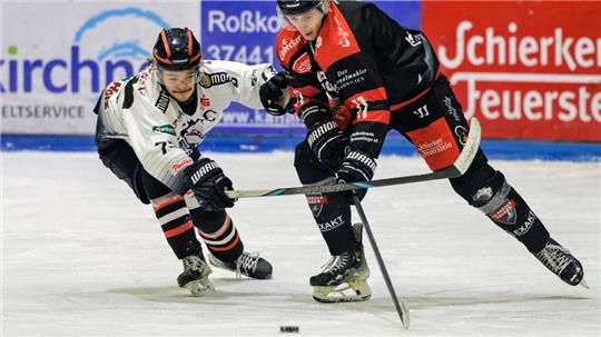 Zwei Eishockeyspieler in Aktion, einer in weißem Trikot, der andere in schwarzem Trikot mit roten Akzenten, kämpfen um den Puck auf dem Eis.