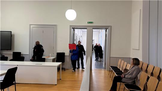 Gerichtssaal mit Stuhlreihe rechts und geöffneter Tür im Mittelpunkt, und einem großen weißen Tisch links in einem Raum mit Holzboden und weißen Wänden. Mehrere Personen kommen zur Tür herein. Eine davon, die im Zentrum steht, hält sich eine rote Mappe vors Gesicht. 