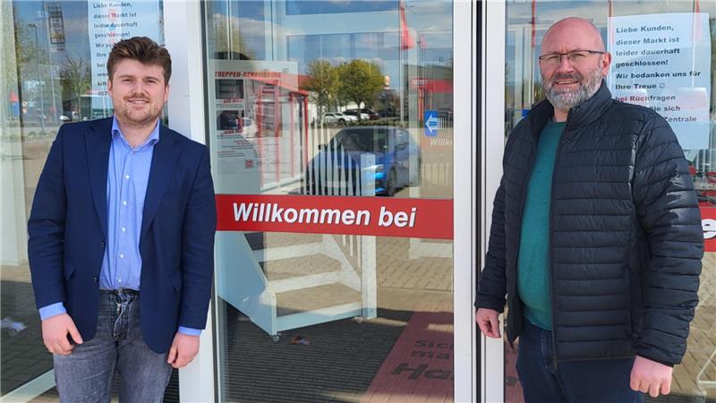 Zwei Männer stehen vor einem Glaseingang mit rotem Schild 'Willkommen bei'