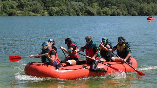 Sechs Personen mit Schwimmwesten paddeln in einem roten Schlauchboot auf einem See