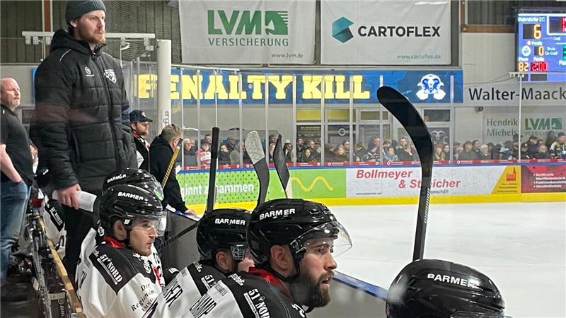 Zahlreiche Eishockeyspieler in weißen Trikot sitzen auf einer Bank. Links steht ein Mann mit Mütze.