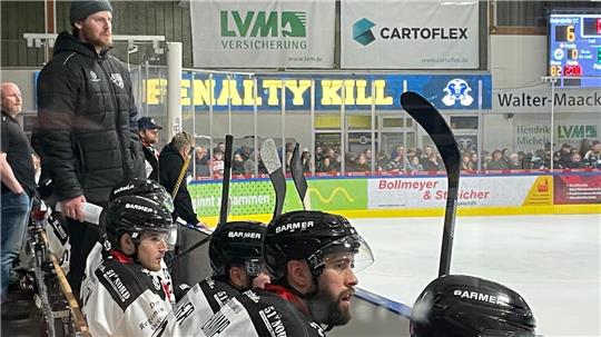 Zahlreiche Eishockeyspieler in weißen Trikot sitzen auf einer Bank. Links steht ein Mann mit Mütze.