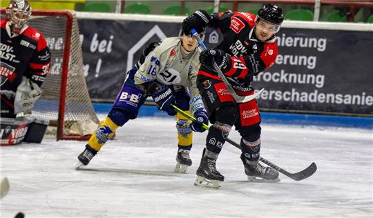 Zwei Eishockeyspieler in Aktion vor dem Tor, einer in weiß-blauem Trikot und einer in schwarz-rotem Trikot, auf dem Eis mit Torwart im Hintergrund