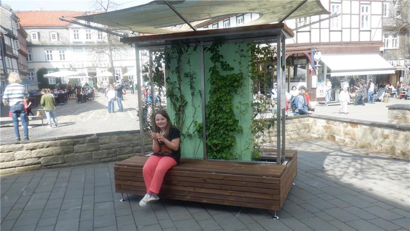 Ein Mädchen sitzt auf einer Holzbank mit grün bewachsenen Seitenwänden in einem belebten Innenhof