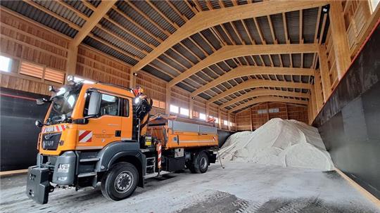 Oranger Lastwagen steht in einer großen Halle neben einem hohen Streusalzhaufen unter einer Holzdecke mit sichtbaren Balken.