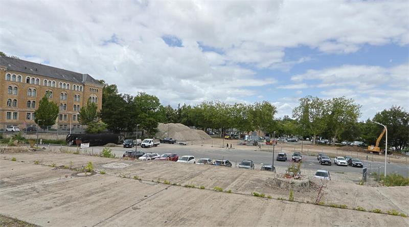 Großes, offenes Baugelände mit mehreren geparkten Autos, einem großen Erdhaufen und einem historischen Backsteingebäude im Hintergrund