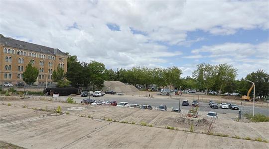 Großes, offenes Baugelände mit mehreren geparkten Autos, einem großen Erdhaufen und einem historischen Backsteingebäude im Hintergrund
