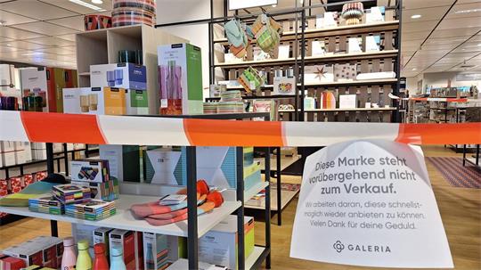 Ein rot-weißes Absperrband hängt vor einem Tisch mit Geschirr in einer Galeria-Filiale. An dem Band hängt eine Notiz: „Diese Marke steht vorübergehend nicht zum Verkauf.“