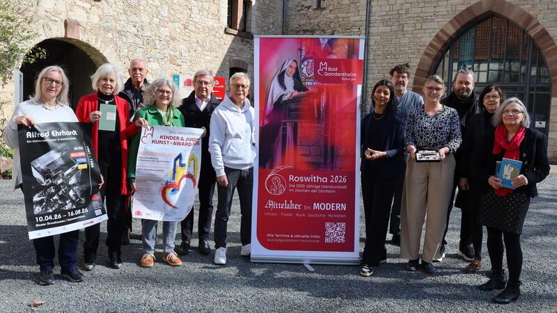 Gruppe von Menschen steht vor einer Steinmauer und hält Plakate und Flyer, daneben ein rotes Werbebanner mit Text und Bild einer Frau