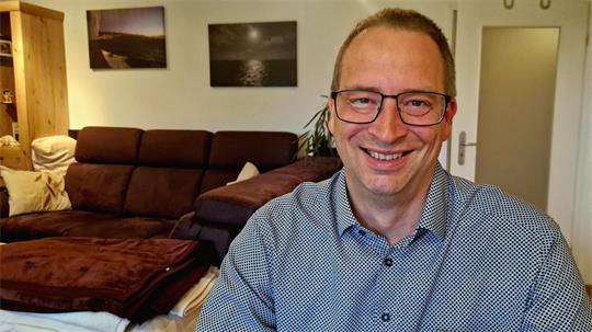 Mann mit Brille sitzt lächelnd vor einem Sofa in einem Wohnzimmer mit Bildern an der Wand.
